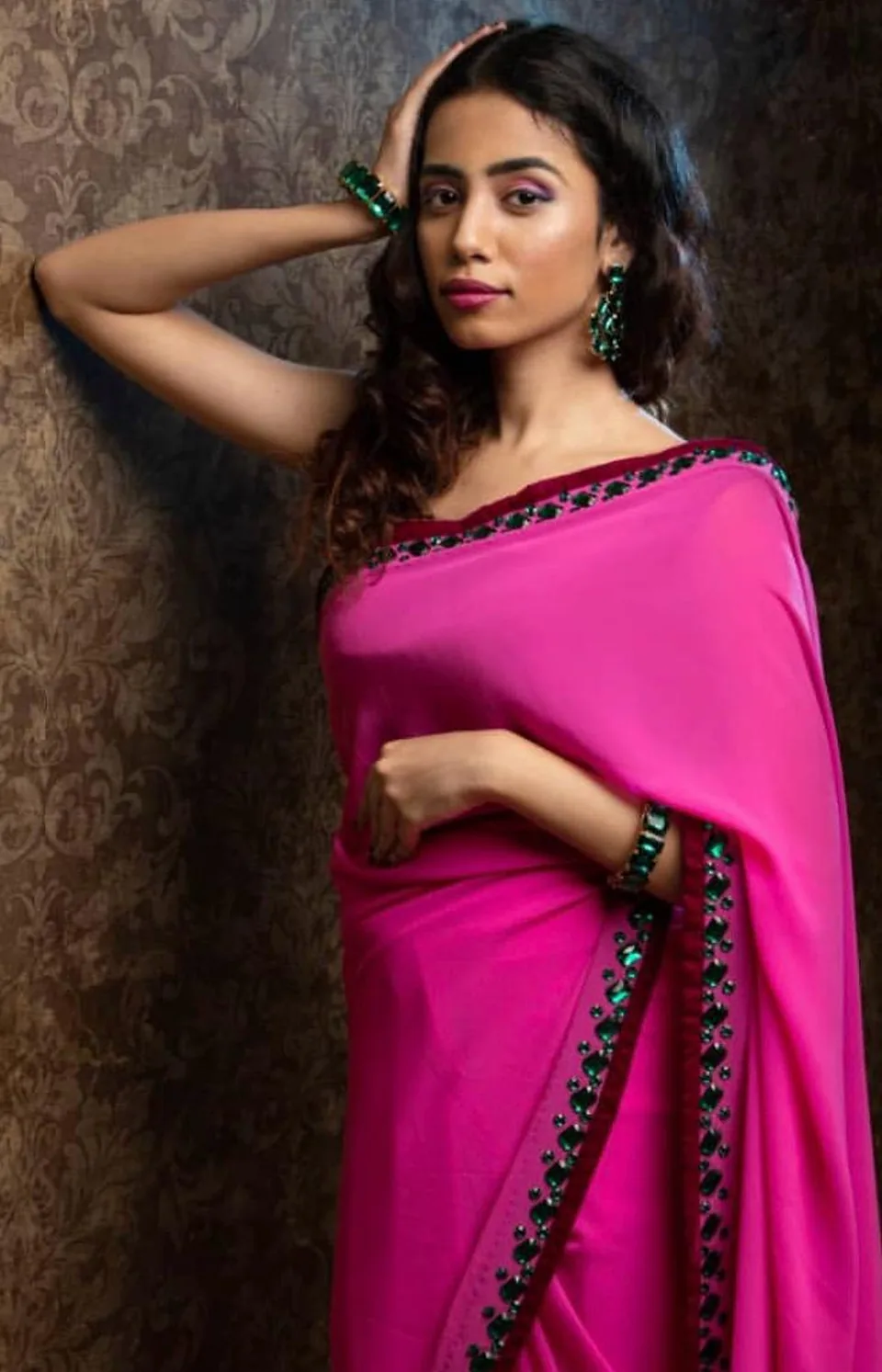Model in the rani pink saree with its emerald stone border, one hand raised to her hair.